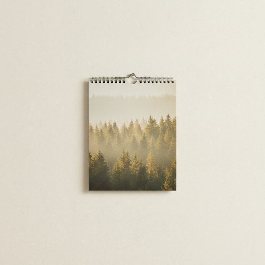 Wandkalender hochkant an heller Wand, Kalenderseite zeigt einen nebligen Kiefernwald im Morgenlicht